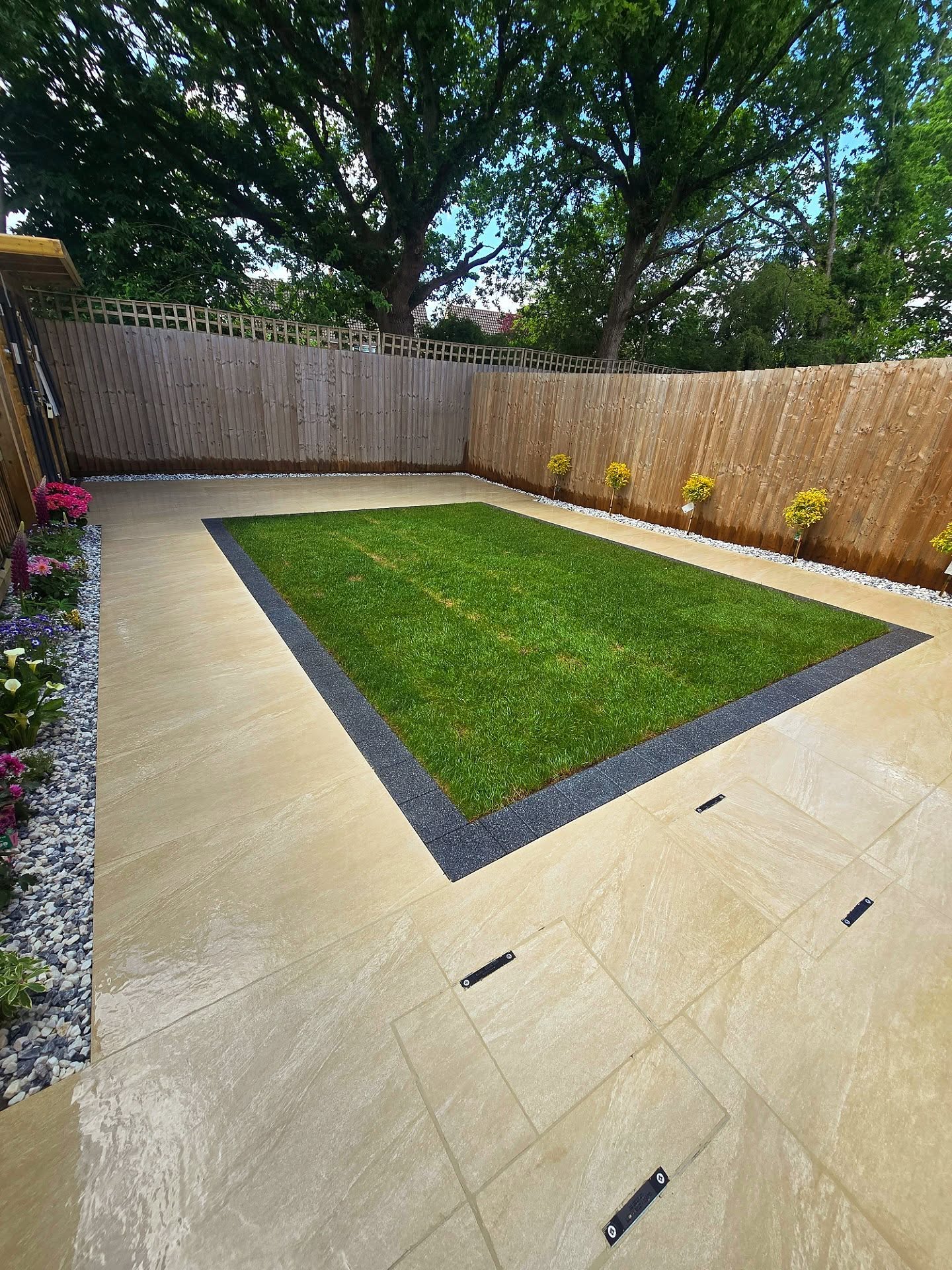 Finished back garden with light-toned paving, a central lawn framed by dark edging and planted borders